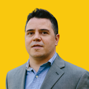 Headshot of Rodrigo Dominguez-Villegas, looking at the camera with a neutral expression, wearing a gray blazer over a light blue button-down shirt against a dandelion-yellow background.