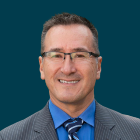 Headshot of Roy Agloinga, smiling and wearing glasses, a gray pinstripe suit, blue shirt and striped tie, against a dark teal background.
