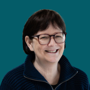Headshot of Sandy Brewster, smiling, with short dark hair and glasses, wearing a dark blue zip-neck sweater against a dark ocean-blue background.