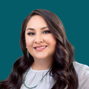 Headshot of Shawna Douma, smiling, with long wavy dark brown hair, wearing a light gray blazer, white blouse, turquoise jewelry and earrings against a dark ocean-blue background.