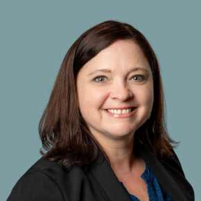 Headshot of Sheila Babb Anderson, smiling, with shoulder-length dark brown hair, wearing a black blazer over a blue patterned blouse against a muted teal background.