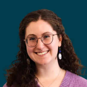 Headshot of Shoshi Bieler, smiling and wearing glasses, dangling earrings and a purple sweater, against a dark teal background.