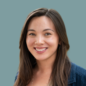 Headshot of Shu-Ling Zhao, smiling, with long brown hair, a small nose ring and delicate jewelry, wearing a blue jacket against a muted teal background.