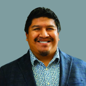 Headshot of Sky Pagaling, smiling, with short dark hair, wearing a blue blazer over a patterned button-down shirt against a muted teal background.
