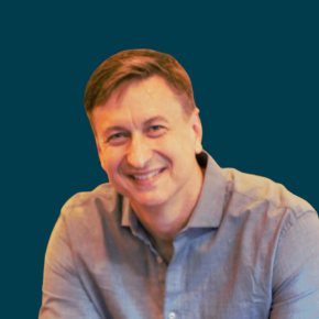 Headshot of Steve Patty, smiling, with short light brown hair, wearing a light blue button-down shirt against a dark ocean-blue background.