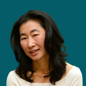 Headshot of Suk Rhee, looking at the camera with a slight smile, with shoulder-length black hair, wearing a cream-colored top against a dark ocean-blue background.