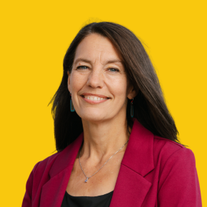 Headshot of Suzanne LaFrance, smiling, with long dark hair, wearing a magenta blazer and beaded earrings against a dandelion-yellow background.