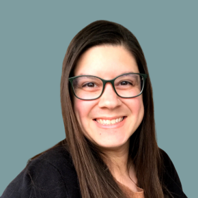 Headshot of Tara Havlicek, smiling, with long straight dark hair and dark-framed glasses, wearing a black cardigan over a tan top against a light grey background.