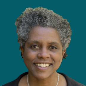 Headshot of Tonisha Toler, smiling, with short natural gray curls, wearing a black top and silver necklace against a dark ocean-blue background.