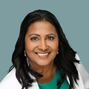 Headshot of Vandana Ingle, smiling with long dark hair and wearing a green top, white blazer and earrings, against a muted blue-gray background.