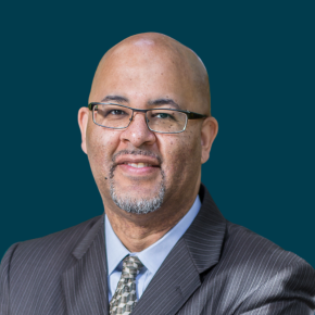 Headshot of Warren Brown, smiling slightly, wearing glasses, a gray pinstripe suit jacket, light blue shirt and patterned tie against a dark ocean-blue background.