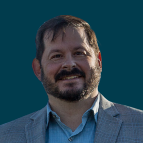 Headshot of Wayne Ducheneaux II, smiling, with short dark hair and a beard, wearing a light gray blazer over a blue shirt against a dark ocean-blue background.