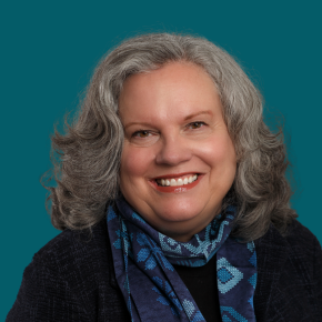 Headshot of Wendy Schneider, smiling, with shoulder-length curly gray hair, wearing a navy jacket and a blue patterned scarf against a dark ocean-blue background.