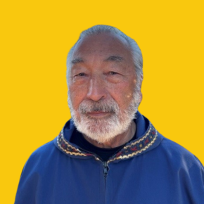 Headshot of William Iggiagruk Hensley, looking at the camera with a slight smile, with white hair and a white beard, wearing a blue zip-up sweater against a dandelion-yellow background.