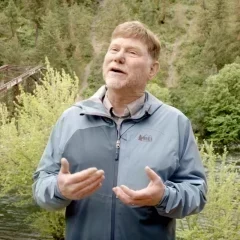 Photo of Leroy Eadie speaking outdoors near a forested river, wearing a light blue-gray jacket with a bridge and green trees in the background.