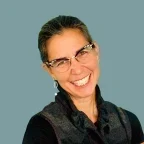 Headshot of Laurie Wolf, smiling, with dark hair pulled back, wearing patterned glasses, dangling earrings, and a dark textured vest against a muted teal background.
