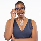Robin Martin, President and CEO of Navigating Courage wearing a sleeveless dark V-neck top and a chunky geometric necklace, posed against a plain white background with one arm crossed and the other raised holding the edge of her glasses.