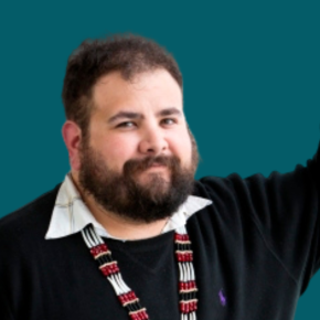 Headshot of Aaron Leggett, with short dark hair and a beard, wearing a black sweater, white collared shirt and beaded necklace, with one arm raised against a teal background.