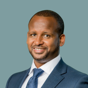 Headshot of Habtamu Bekele, smiling, wearing a blue suit jacket, white dress shirt and blue patterned tie against a muted teal background.