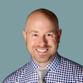 Headshot of Joseph Accuardi-Gilliam, smiling with a shaved head and short beard, wearing a blue-and-white checkered shirt and silver tie, against a muted blue-gray background.