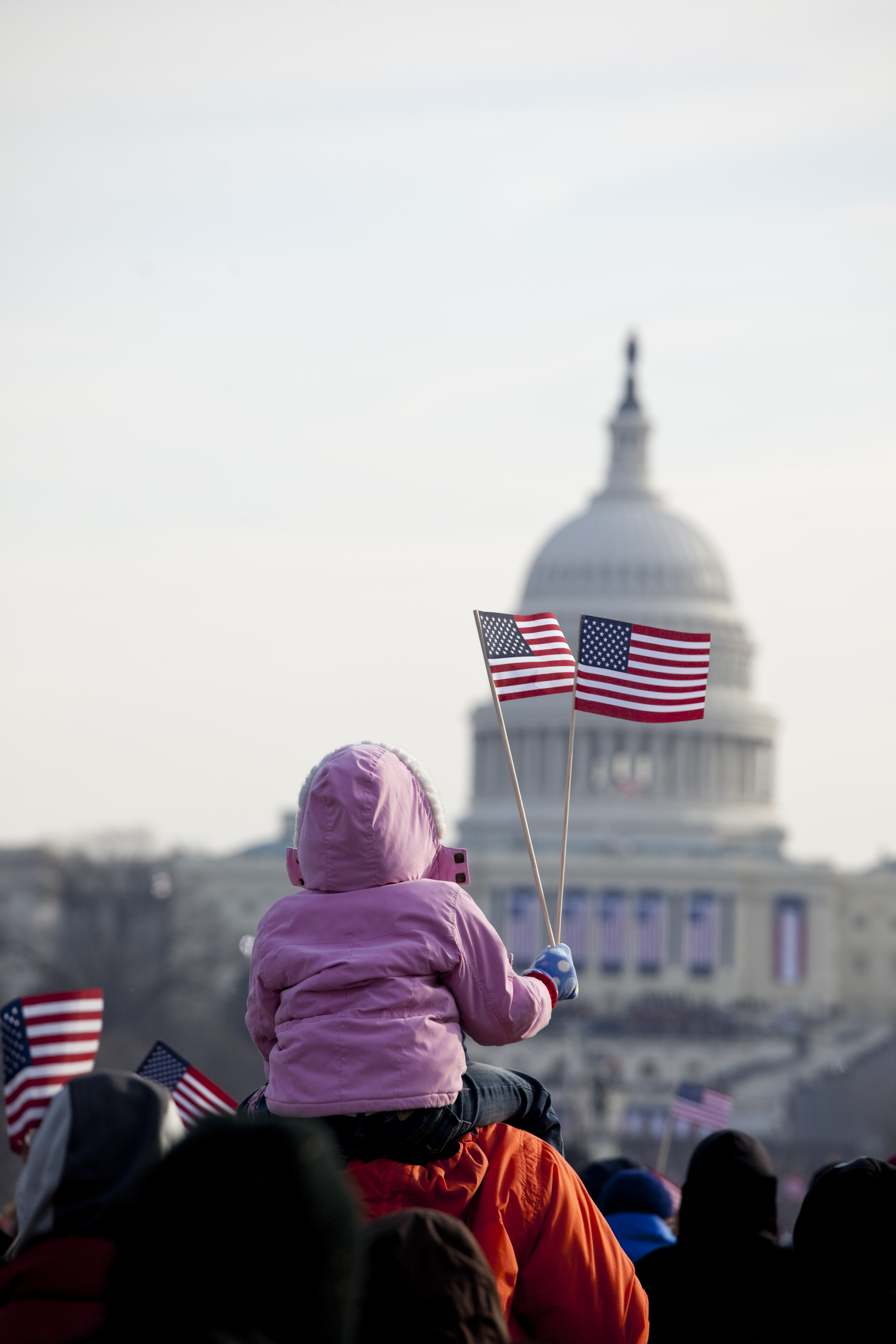 Image of a child on parent's shoulders waving a small US flag in a crowd of people in front of the U.S. capitol