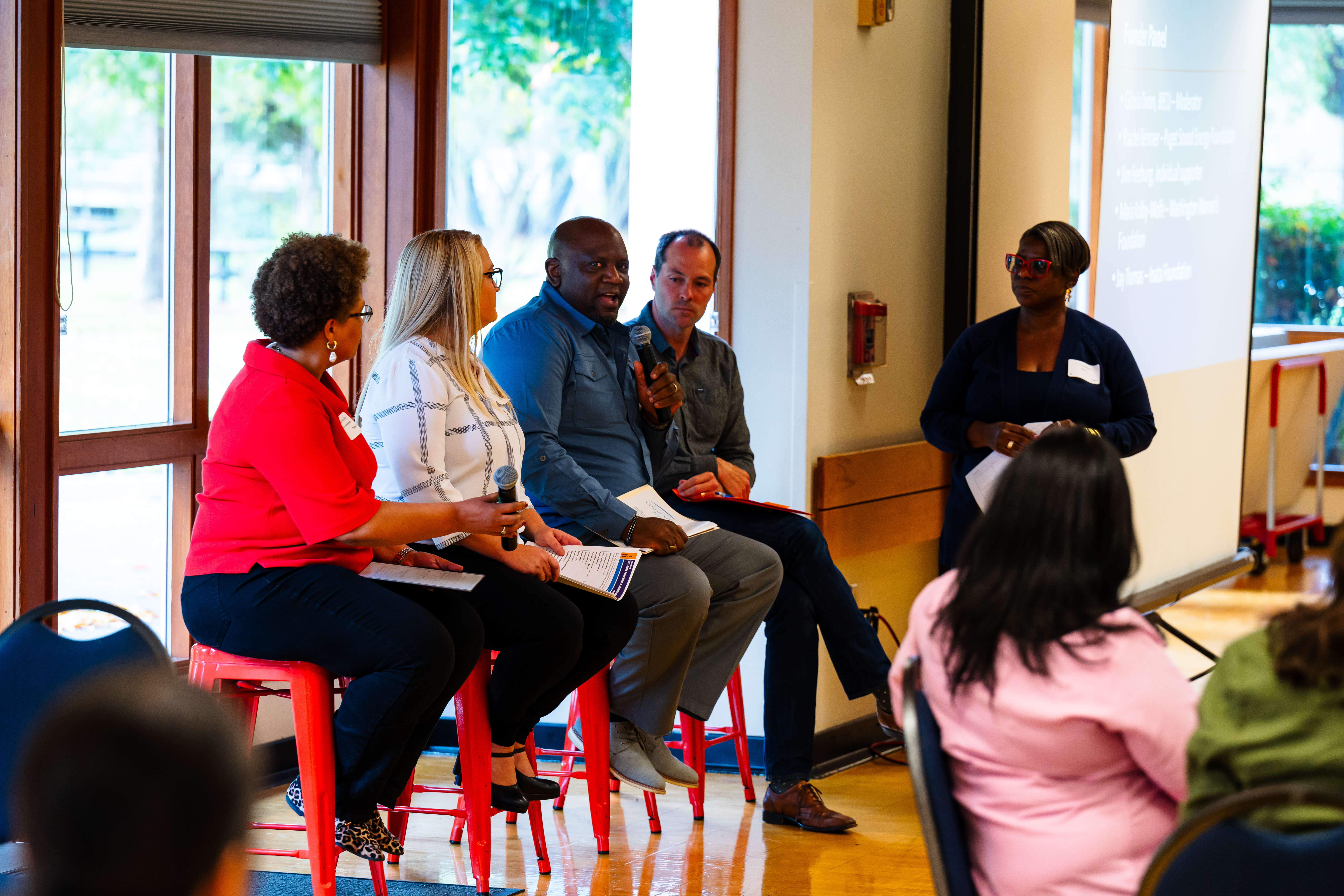 A panel of four speakers sits on red stools in a well-lit room, engaging in a discussion. One speaker holds a microphone while addressing the audience, and the others hold notes and microphones, listening attentively. A moderator stands nearby