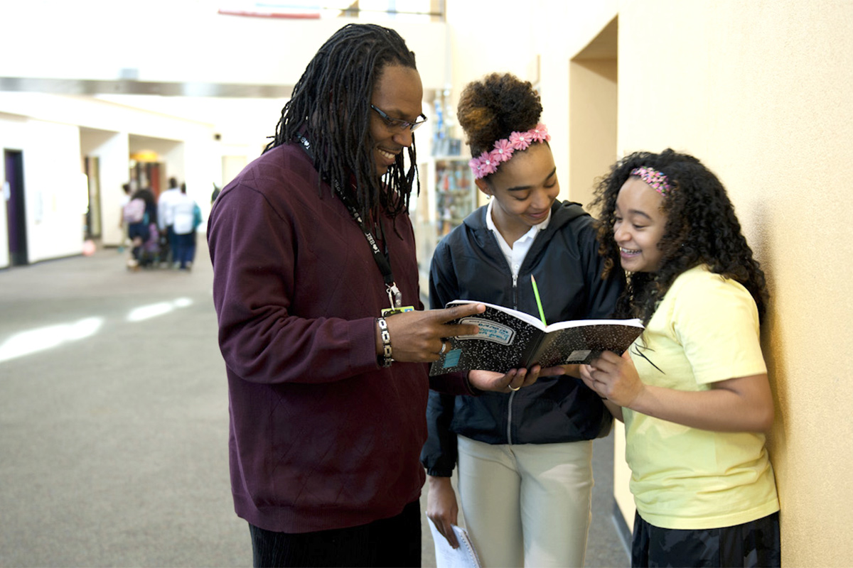 African American male teacher looks at a notebook with two African American teenage girls in school atrium