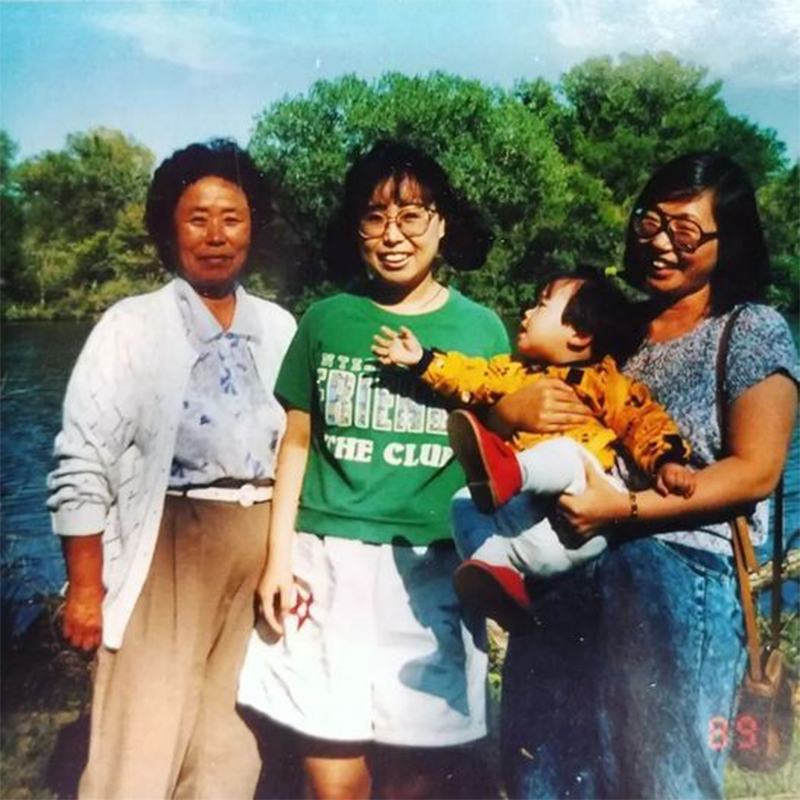 Three adult women and a toddler stand beside a lake with trees behind them. The left woman wears a white cardigan and beige pants; the center woman wears a green T-shirt and white skirt; the right woman holds the child in a yellow jacket and red shoes. They smile, suggesting a warm family outing; a small red “89” appears in the lower right.