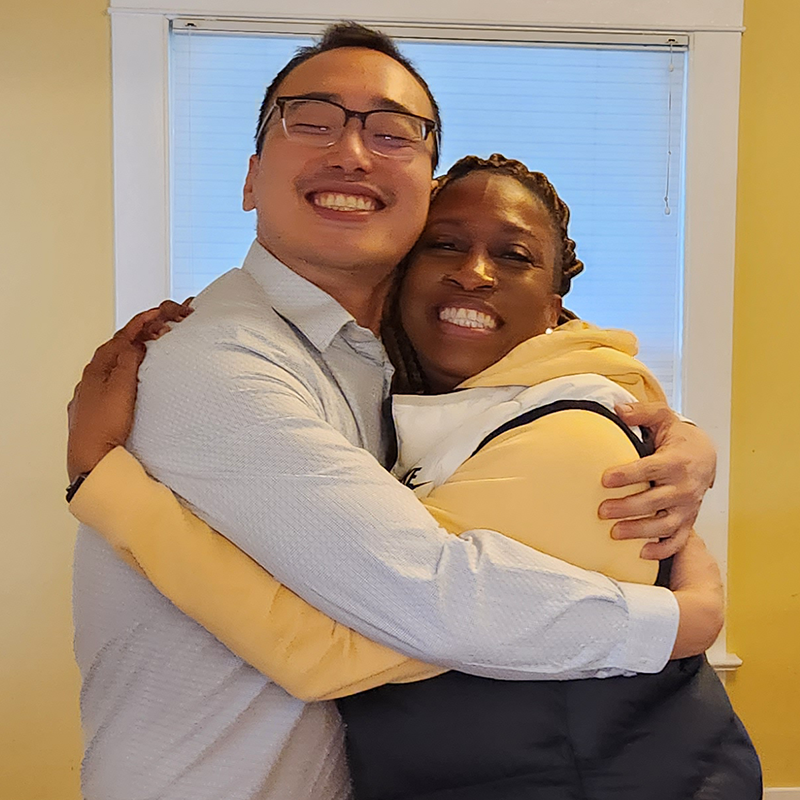 Photo of two people smiling and hugging indoors in front of a window.