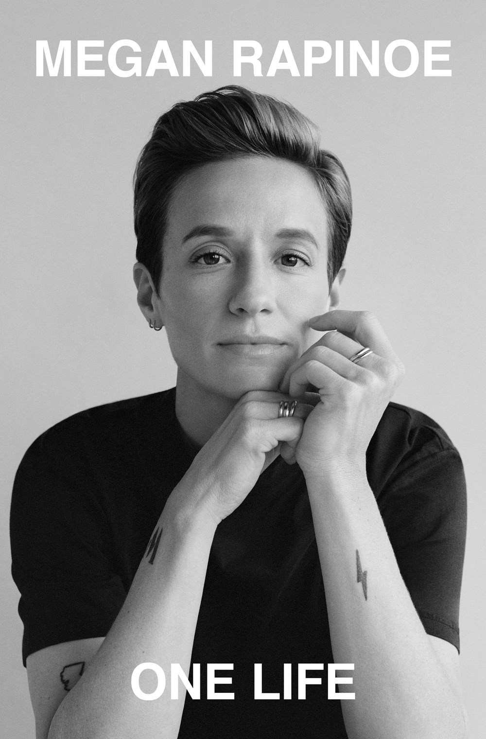 Book cover for “One Life” by Megan Rapinoe, featuring a black-and-white portrait of Rapinoe resting her chin on her hands, with large white text at the top and bottom.