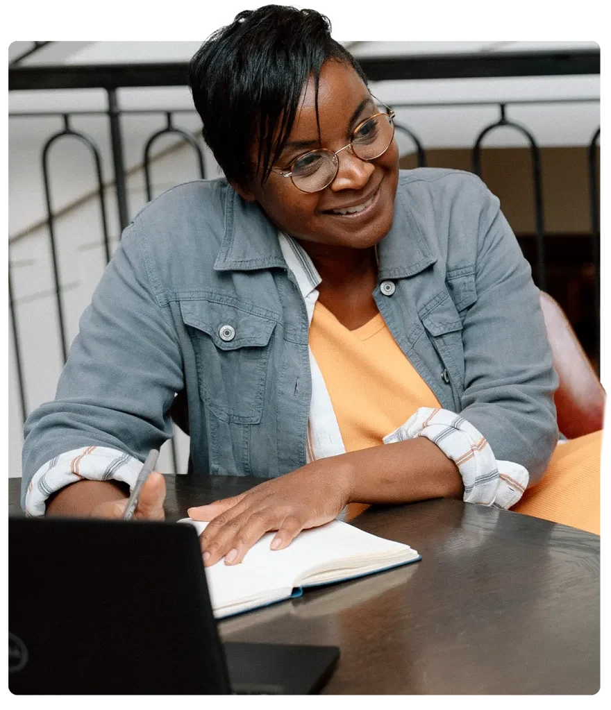 Photograph of TGP Senior Advisor Lisa McGill wearing a denim jacket and light orange desk smiling with a laptop and notebook in front of her.