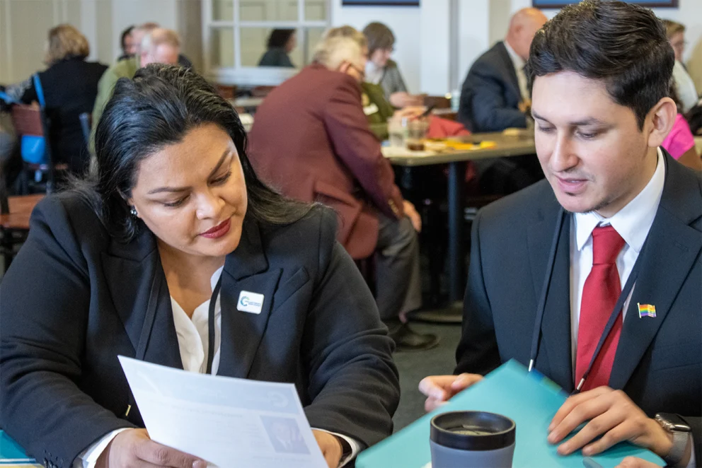 A photo of two attendees seated at a table during Foundations on the Hill, reviewing documents and holding folders. A coffee cup is on the table in front of them. Other participants are visible in the background, engaged in conversation around tables.