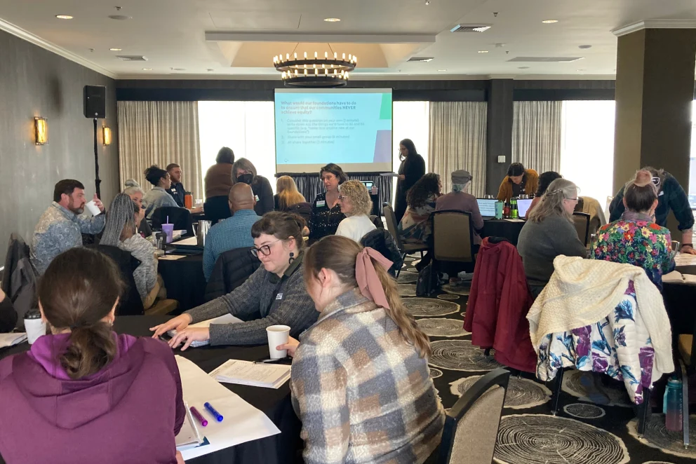 Participants seated at round tables in a conference room during Philanthropy Northwest’s Grantmaking with an Equity Lens (GEL) workshop in Eugene, Oregon. A presentation slide is projected at the front of the room, and attendees are engaged in discussion with papers, notebooks, and drinks on the tables. The space features dark walls, patterned carpet, and a chandelier overhead.