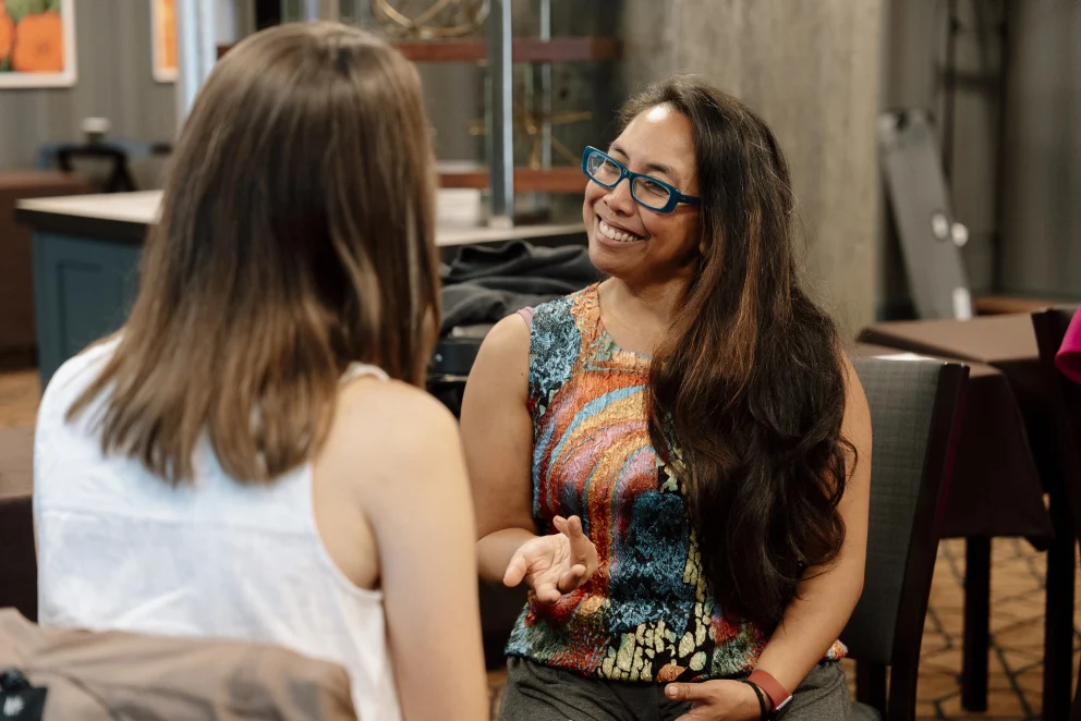 TGP senior advisor Abby engaging in conversation. She sits smiling and leaning forward, wearing blue glasses and a colorful patterned top, with long dark hair over one shoulder. She speaks with another adult seated across from her, shown from behind in a white sleeveless shirt. The setting appears to be an informal meeting space, conveying warmth, attentiveness, and connection.
