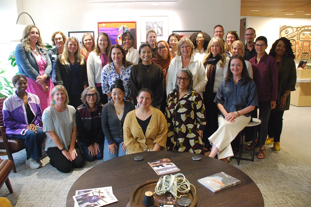 Group photo of Philanthropy Northwest’s Regional Community Philanthropy Group during their fall 2025 convening in Seattle, with about 25 people standing and sitting in a meeting space. A round table with magazines and a decorative item is in the foreground; colorful artwork hangs on the wall behind the group.