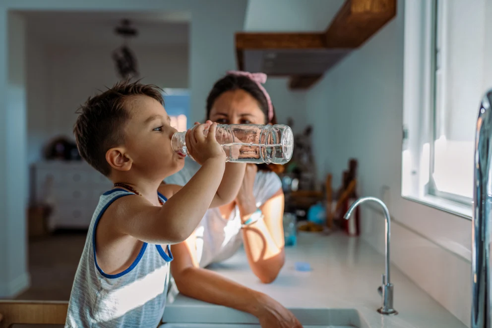 Image of young child drinking water by the kitchen sink with their mother standing next to them.