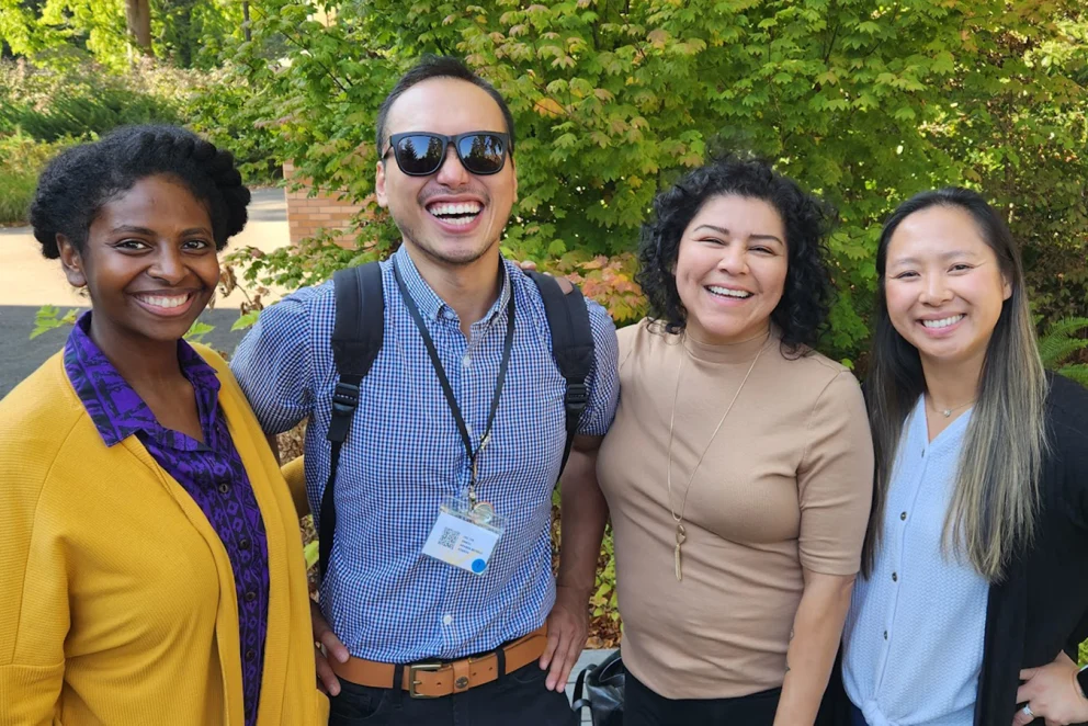 Photo of four people smiling together outdoors in front of green trees.