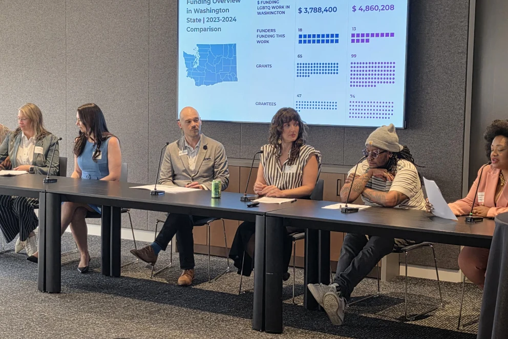 Photo of a panel discussion in a meeting room, with seven speakers seated behind microphones at a long table. A presentation slide behind them shows a funding overview in Washington state.