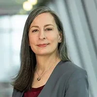 Headshot of Cat Martin miling and wearing a gray blazer, with a softly blurred indoor background of windows and lights.