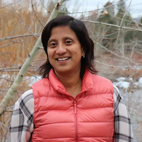 Headshot of Brenda Solórzano, smiling outdoors, wearing a coral puffer vest over a plaid shirt with bare tree branches in the background.