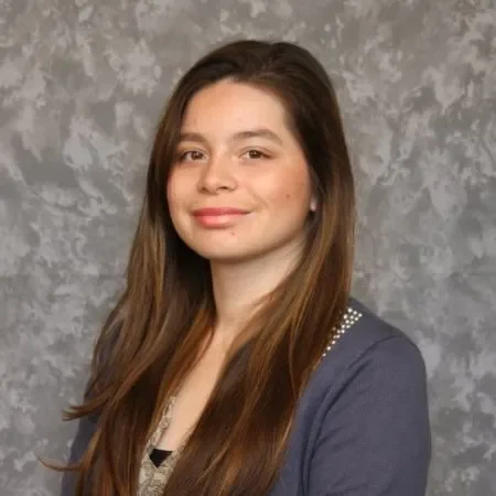 Headshot of Lilliane Ballesteros, looking at the camera with a slight smile, with long brown hair, wearing a navy cardigan against a gray studio backdrop.