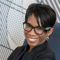 Headshot of Toya Randall, smiling, with short black hair and glasses, wearing a black jacket against an abstract patterned background.