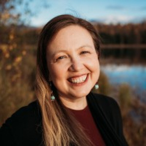 Headshot of Aleesha Towns-Bain smiling, wearing turquoise earrings and a dark jacket, with a lakeside landscape and autumn trees in the background.