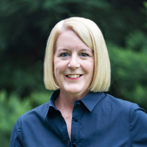 Headshot of Beth McCaw smiling, wearing a navy collared shirt, with a blurred background of green trees.