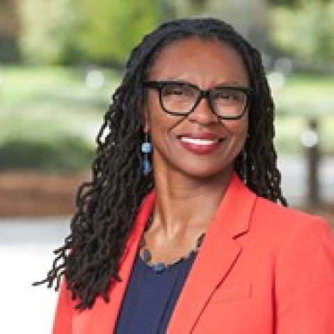 Headshot of Charmaine Mercer smiling, wearing glasses, a coral blazer and a dark top, with a blurred outdoor background of trees and greenery.