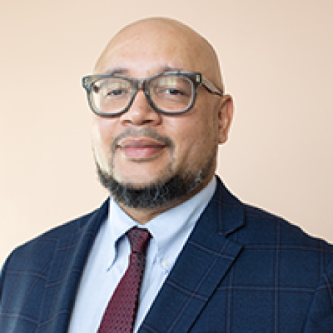 Headshot of D’Artagnan Caliman smiling, wearing glasses, a checked navy blazer, light blue shirt and burgundy tie against a soft peach background.