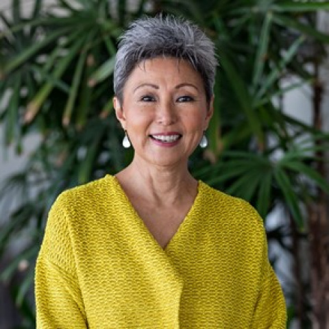 Headshot of Debra Nakatomi smiling, wearing a bright yellow textured top and pearl earrings, with tall green plants in the background.