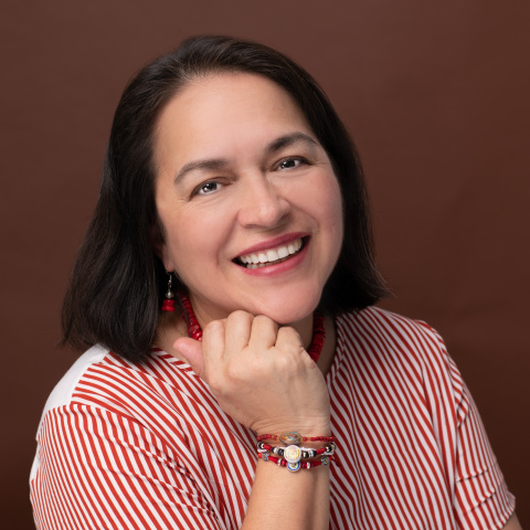 Headshot of Dilcia Molina-Sanchez smiling, resting her chin on her hand, wearing a red-and-white striped top and beaded jewelry, against a solid brown background.