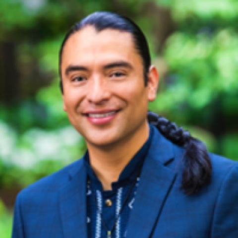 Headshot of Estakio Beltran smiling, wearing a blue blazer and patterned shirt, with a lush green outdoor background.