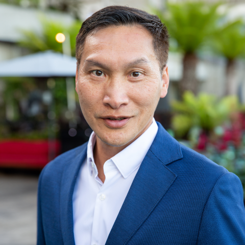 Headshot of Eddy Zheng smiling, wearing a blue blazer and white shirt, with an outdoor setting of plants and blurred seating in the background.